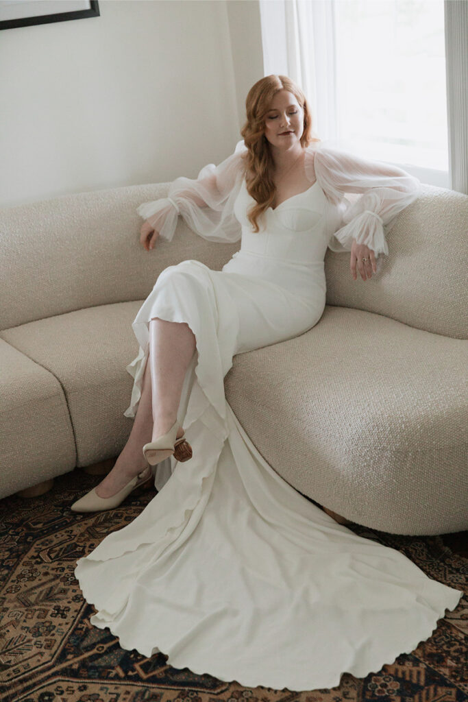 Person in a white dress with sheer sleeves sits on a light-colored couch, with a long train draped over a patterned rug.