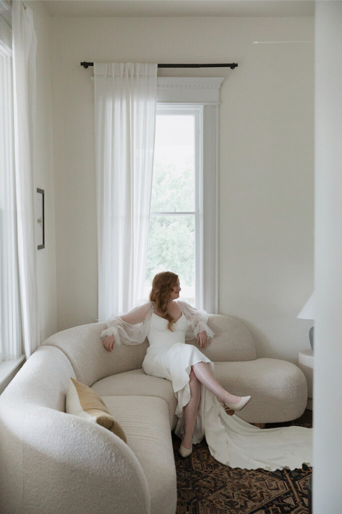 Woman in a white dress sits on a beige couch by a window of The Grand Lady Austin with sheer curtains, looking to the side.