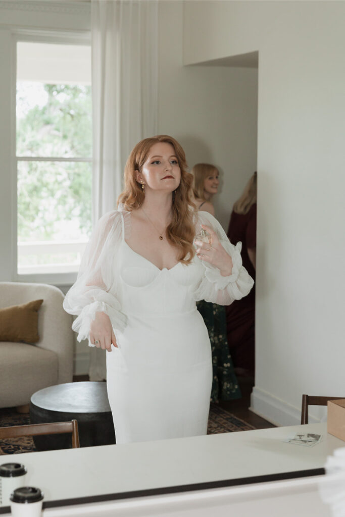 Woman in a white dress stands indoors holding a wine glass, with another person in the background. The room has a large window and a sofa.