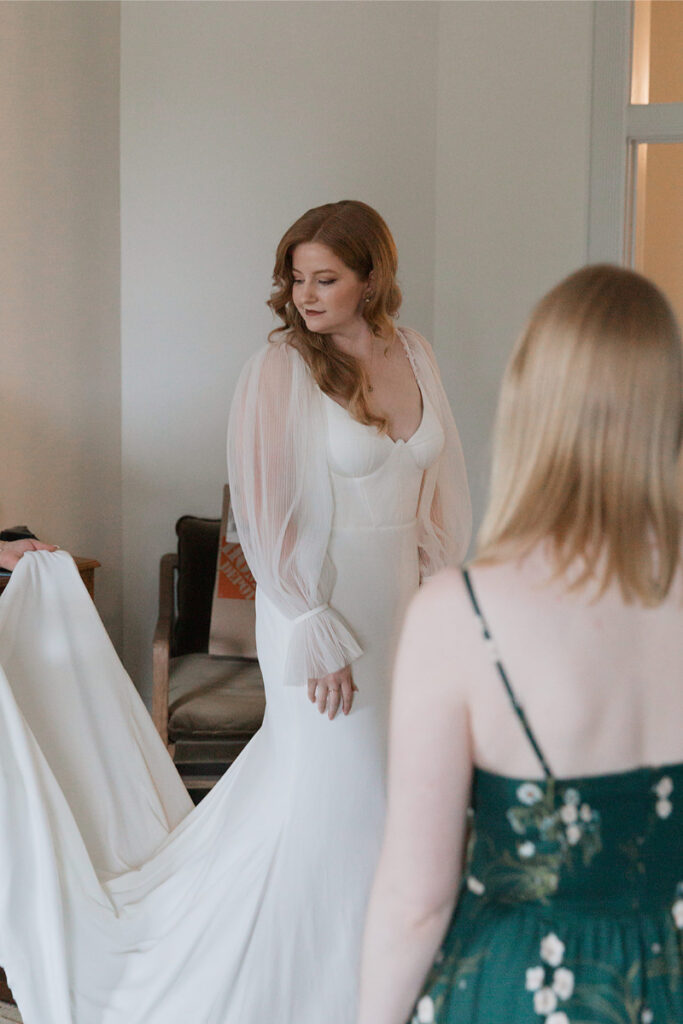 A bride in a white wedding dress with sheer sleeves is assisted by another woman in a floral dress as they prepare indoors.