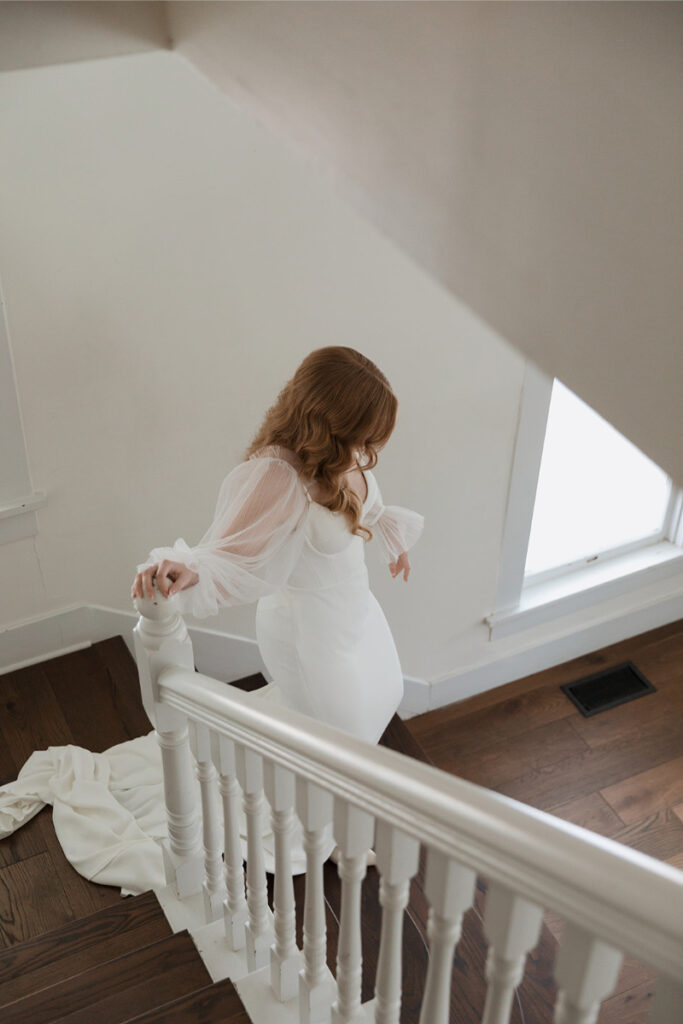 A woman in a white gown descends a wooden staircase of The Grand Lady Austin. She has long, wavy hair and is holding part of her dress. The staircase has white railings.