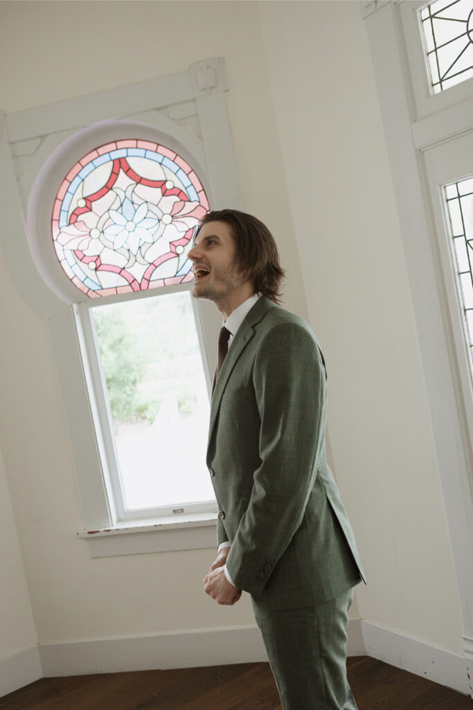 A man in a green suit stands in a room with stained glass windows, looking upward and appearing to speak or sing.