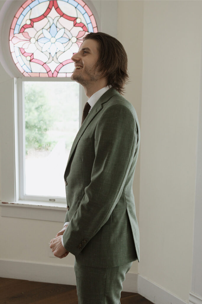 A man in a green suit stands indoors near a stained glass window, smiling.