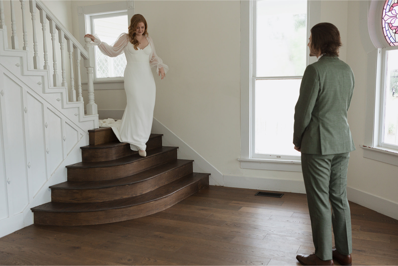 A person in a white dress descends a staircase of The Grand Lady Austin while another in a green suit stands watching in a room with wooden floors and large windows.