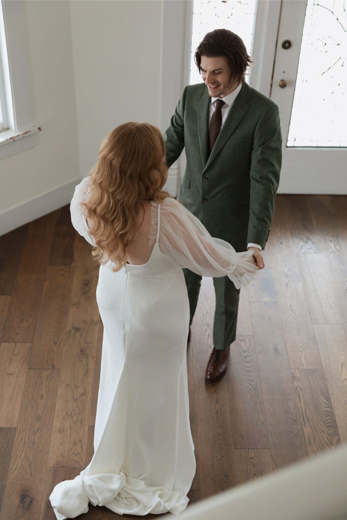 A couple is inside, holding hands. The woman wears a white wedding dress, and the man wears a green suit and tie. They stand on a wooden floor near a window and a door.