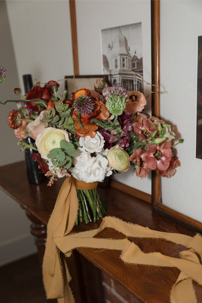 A bouquet of mixed flowers with greenery and long beige ribbons sits on a wooden mantel against framed pictures.