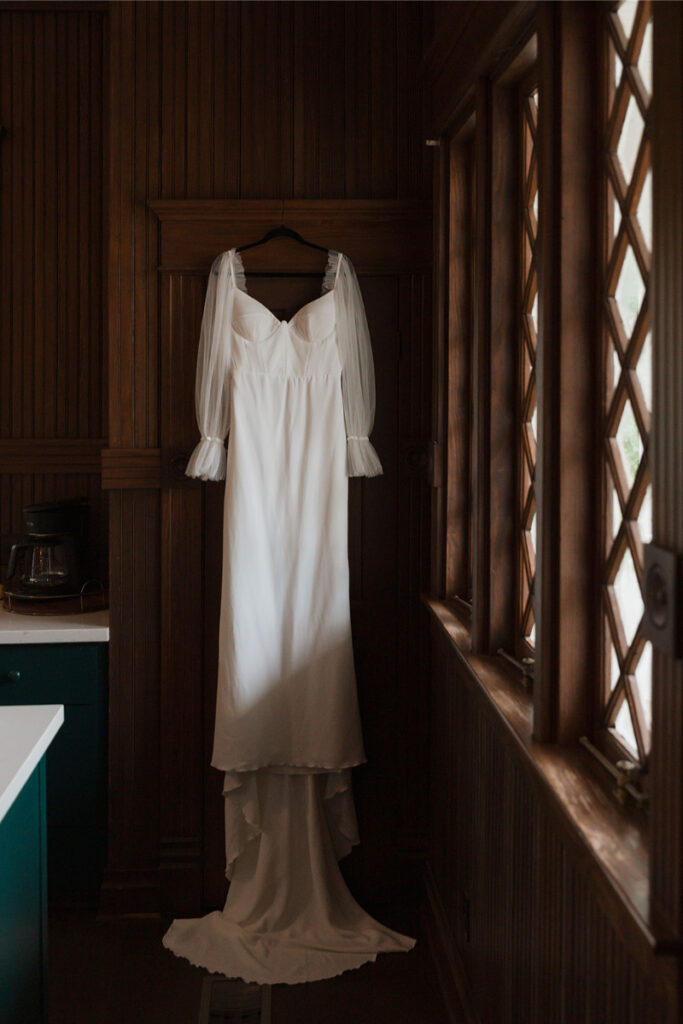 A white dress with sheer long sleeves hangs on a black hanger in a wooden-paneled room with a diamond-patterned window.