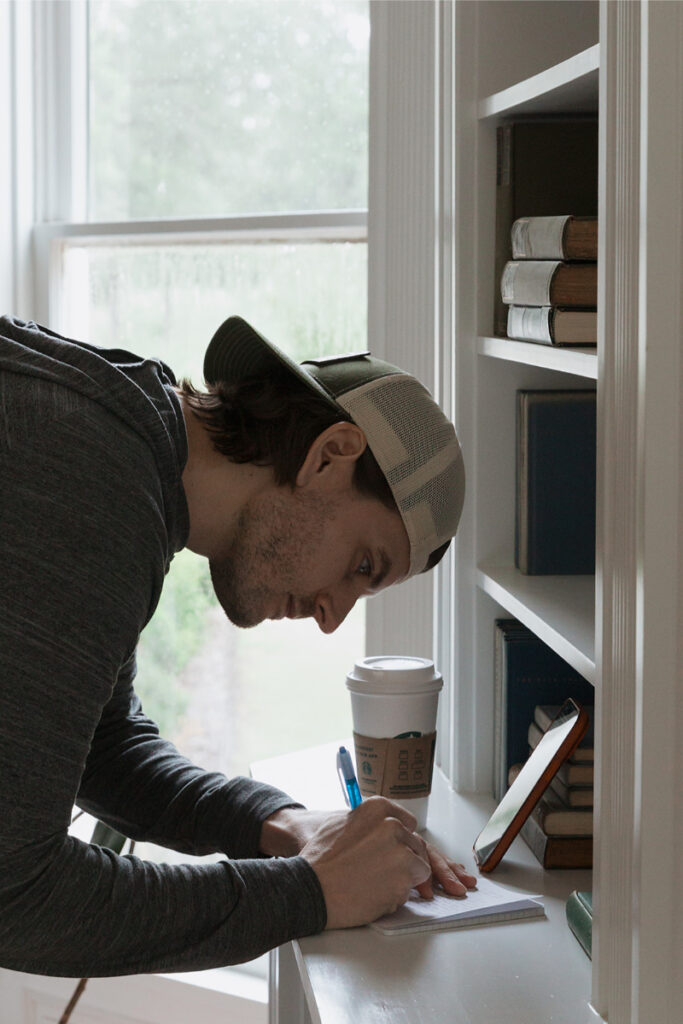 Man in a cap writing in a notebook by a bookshelf with books and a coffee cup nearby.