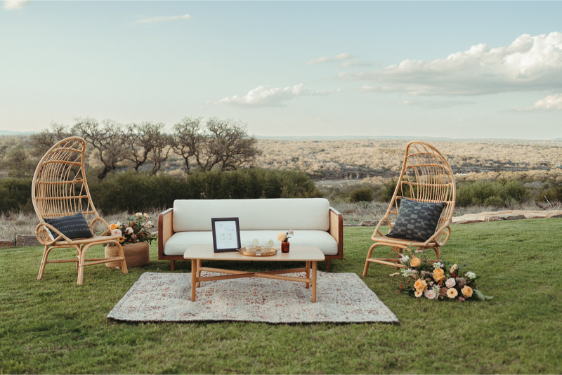 Outdoor seating on grass with a sofa and two wicker chairs around a small table with decor. Rug underneath, floral arrangements, and landscape with trees in the background.