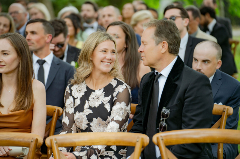 A woman in a floral dress and a man in a suit sit at an outdoor event, smiling at each other.