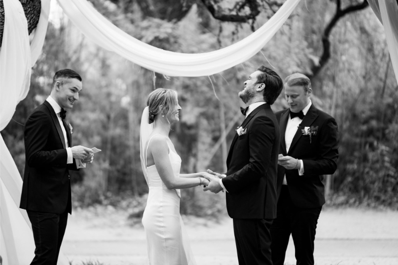 A couple holds hands and smiles at each other during a wedding ceremony, standing under draped fabric, with two people in suits nearby.