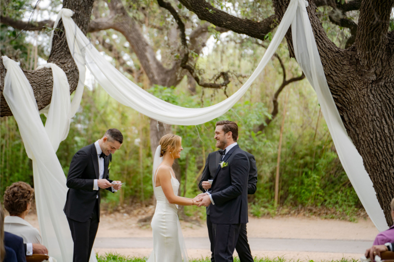 A couple holds hands during their outdoor wedding ceremony, with white drapes hung between trees. A man stands nearby reading from a small book.