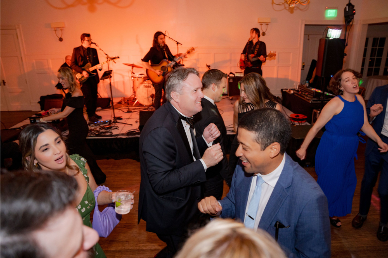 People dancing at an indoor event with a live band performing on stage in the background.