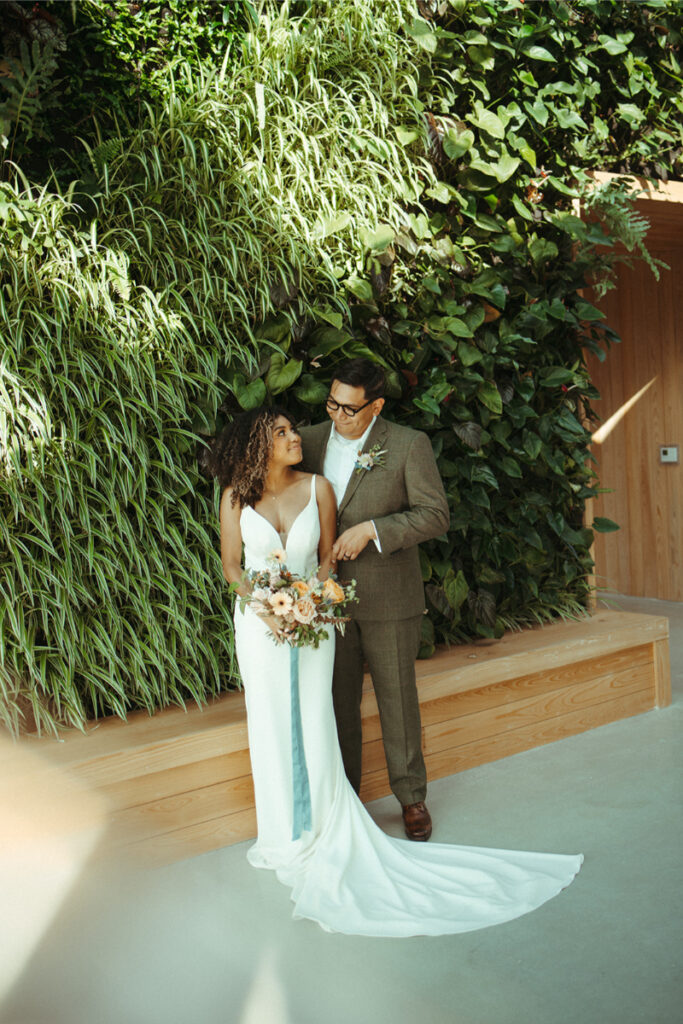 A bride and groom stand together in a bright space with a lush green wall. The bride holds a bouquet, and both are looking at each other.