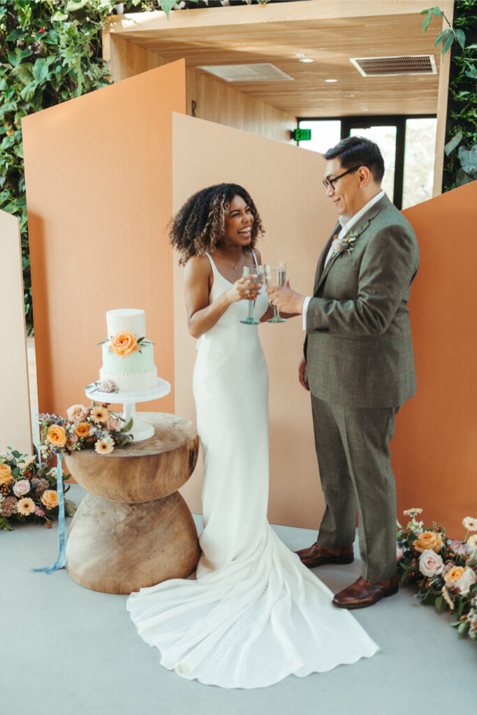 A bride and groom toast with champagne glasses beside a decorated wedding cake on a wooden stand, surrounded by flowers, happy that In Ink Weddings helped them with their wedding planning checklist.