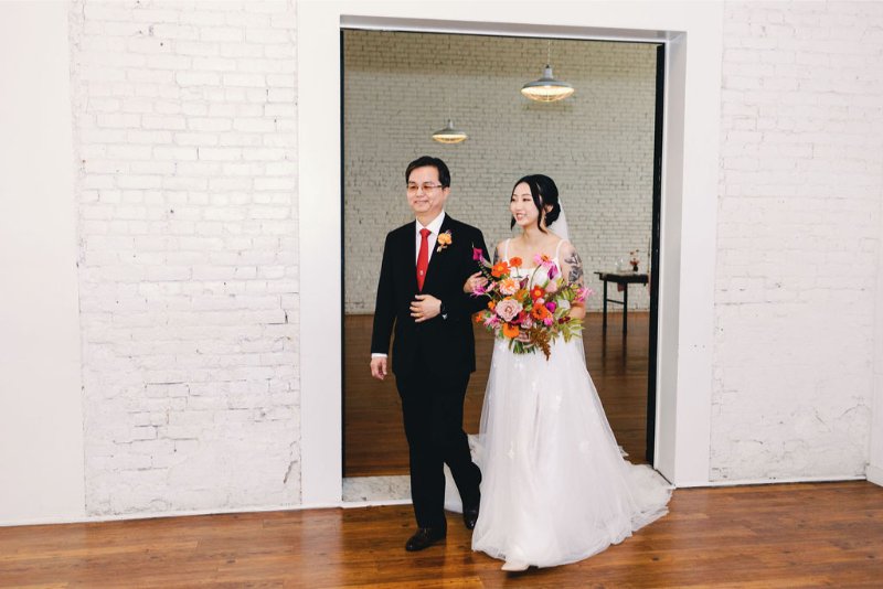 A bride in a white gown holding a bouquet walks arm-in-arm with father in a suit and red tie through a doorway into One Eleven East Hutto, entering a room with white brick walls and wooden floors.