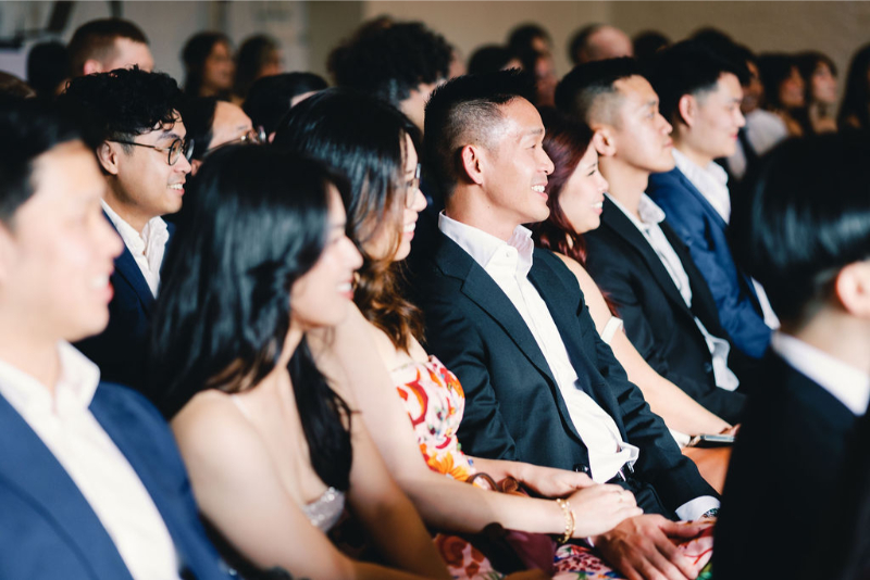 A group of people dressed in formal attire sit closely together in rows at One Eleven East Hutto, attentively watching an event.