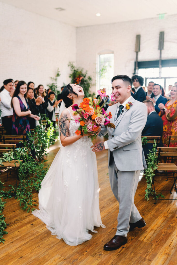 A bride and groom walk down the aisle smiling, holding a colorful bouquet, with guests clapping among lush greenery at the bright indoor venue of One Eleven East Hutto.
