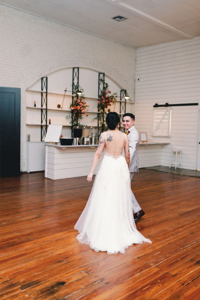 A bride in a white dress and a groom in a light suit dance together on the polished wooden floor at One Eleven East Hutto, surrounded by white walls and elegant floral decorations.