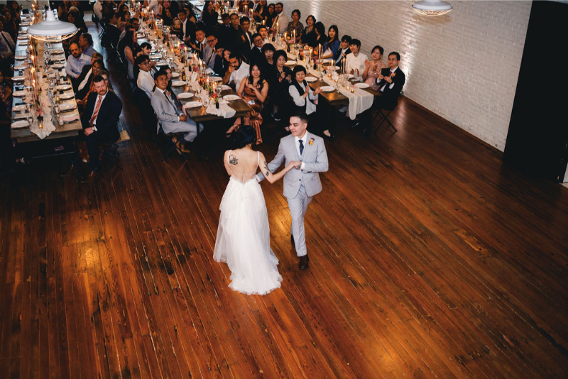 A bride and groom dance on a wooden floor at One Eleven East Hutto while wedding guests seated at long tables watch from the sides.