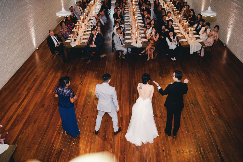 A bride and groom stand with two others at the front of a large reception hall at One Eleven East Hutto, addressing seated guests at long, candlelit tables.