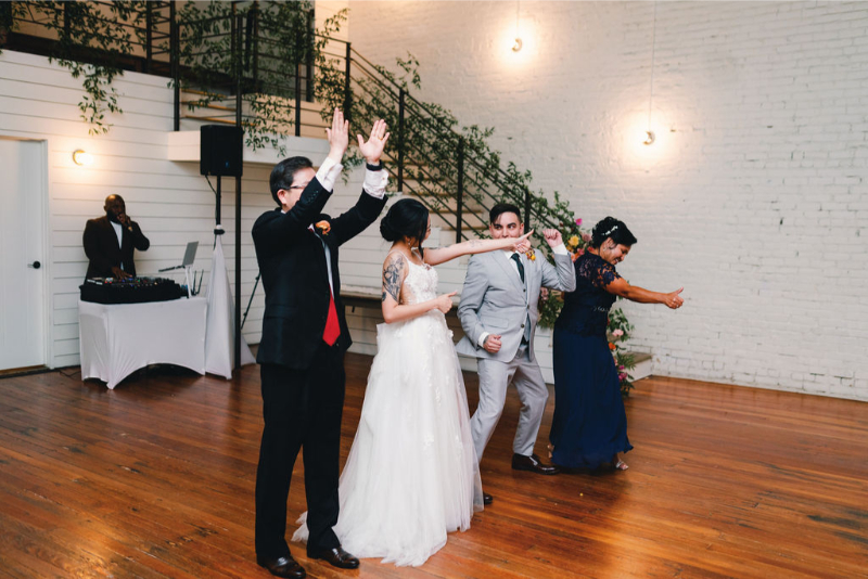 Four people dressed formally are dancing in a line on a wooden floor at One Eleven East Hutto, in a bright room with a DJ in the background near a sound table.