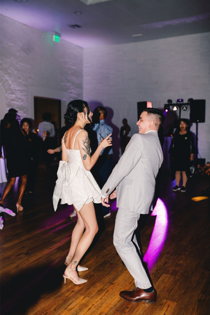 A woman in a short white dress and a man in a light suit dance together on the wooden floor at One Eleven East Hutto, with other people enjoying the indoor event in the background.