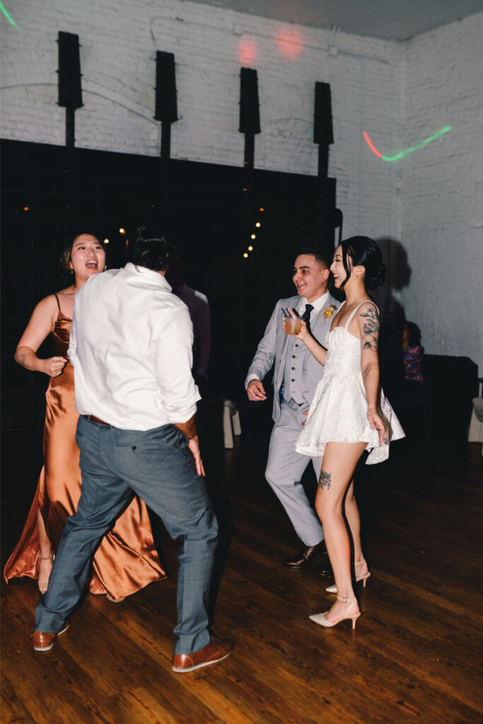 Four people are dancing together on a wooden floor in formal attire at One Eleven East Hutto, with a white brick wall and dark windows in the background.