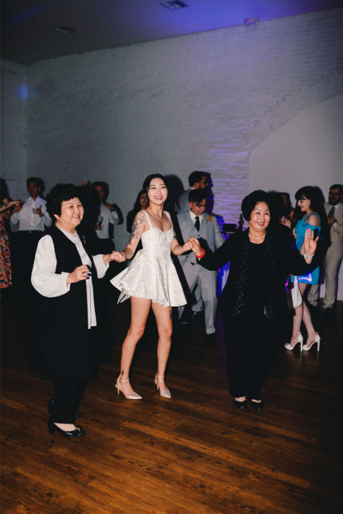 Three women, including a young woman in a short white dress, hold hands and dance together on the wooden floor at One Eleven East Hutto during an indoor event, with other guests mingling in the background.