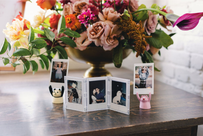 A wooden table holds a floral arrangement in a gold vase, three small photo prints, and two animal-shaped photo holders shaped like a panda and a pig.