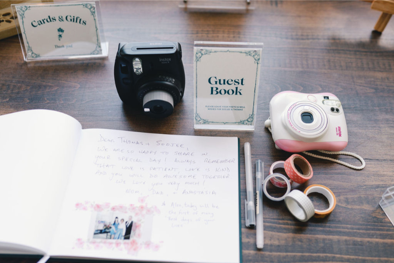 A guest book table with instant cameras, pens, rolls of tape, and a handwritten note, alongside "Cards & Gifts" and "Guest Book" signs.