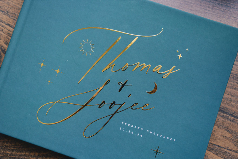 A teal wedding guestbook with gold lettering that reads “Thomas + Joojee," decorated with stars and a moon, and the date 10.26.24 printed at the bottom right.