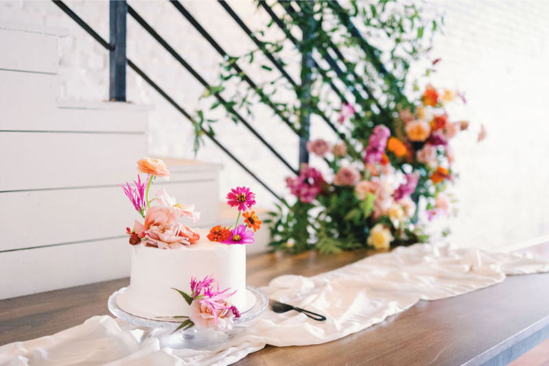 A white frosted cake decorated with pink and orange flowers sits on a wooden table with a white cloth, beside a staircase adorned with more colorful flowers.