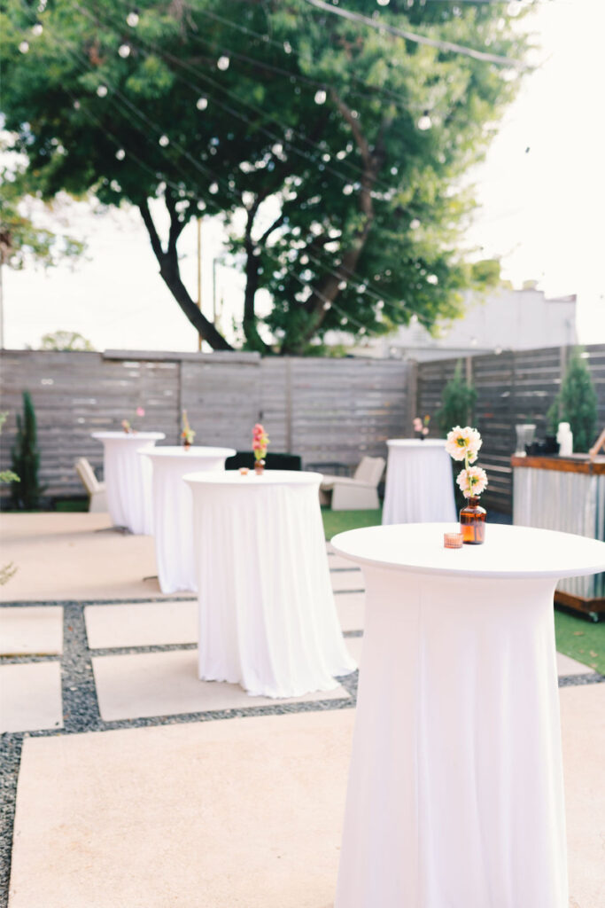 Outdoor event space with tall cocktail tables covered in white tablecloths, each decorated with a small vase of flowers, under string lights and surrounded by a wooden fence.
