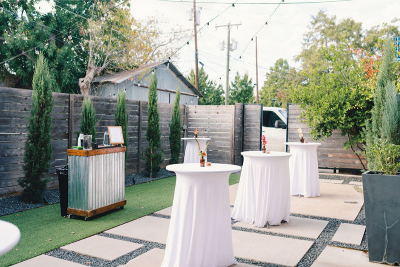 An outdoor event space with cocktail tables covered in white cloths, a small bar on the left, string lights above, and trees along a wooden fence.