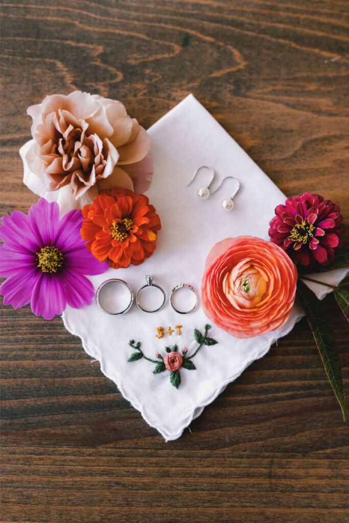 A white embroidered handkerchief with rings, pearl earrings, and five colorful flowers arranged on a wooden surface.