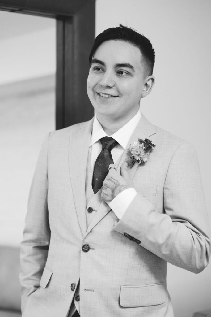 A person in a light-colored suit and tie stands indoors, smiling and adjusting their jacket, with a boutonniere on their lapel.