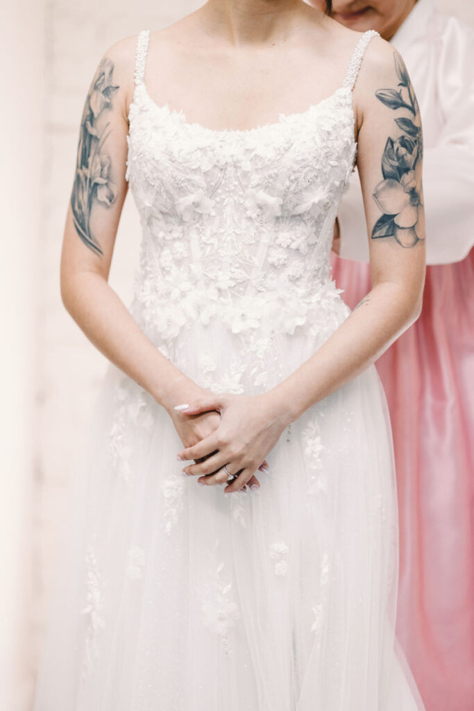 A person with floral arm tattoos wears a white, floral-embroidered wedding dress while another person in pink assists from behind.