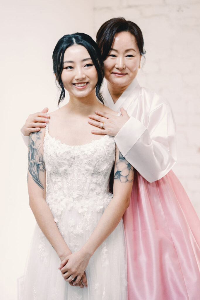 A bride in a white dress stands in front of a woman wearing a pink hanbok, both smiling and posing for a photo against a light background.