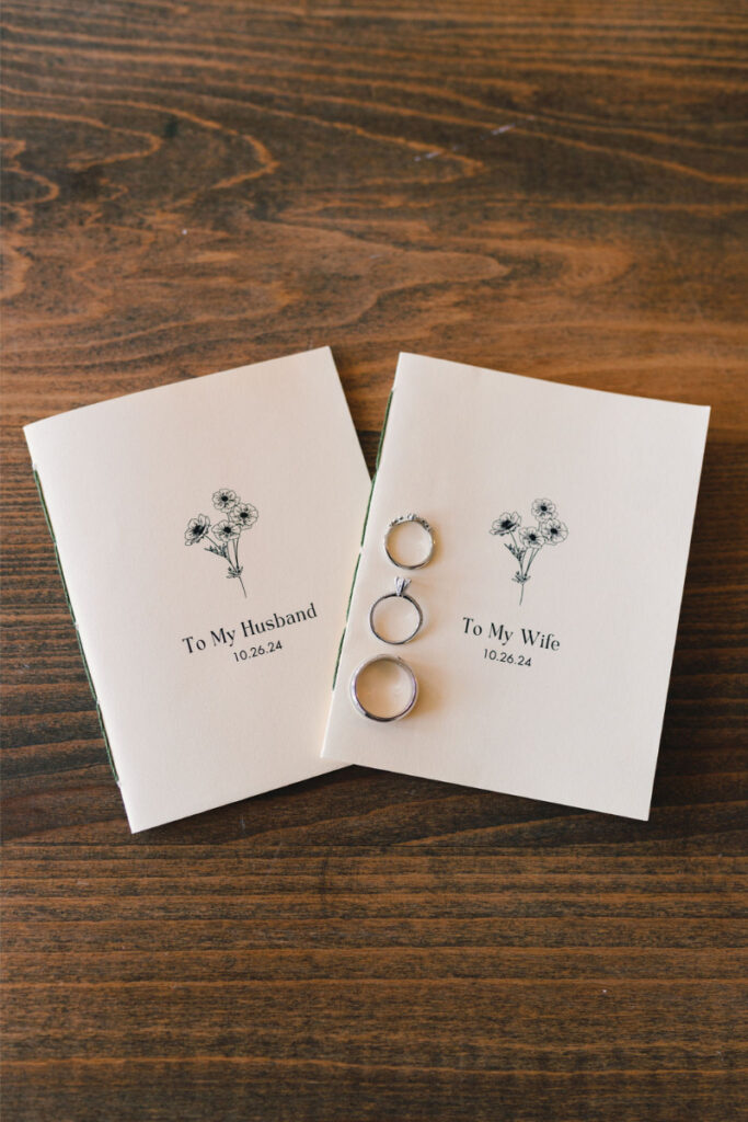 Two cream-colored booklets labeled "To My Husband" and "To My Wife" with illustrated flowers, dated 10.26.24, and three silver rings placed on top, on a wooden surface.