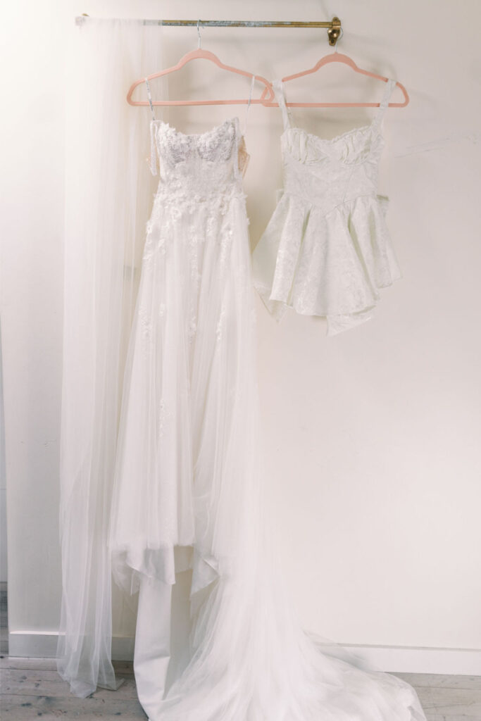 A white wedding dress with a long train and lace details hangs next to a short white corset-style peplum top on a wall-mounted rod.