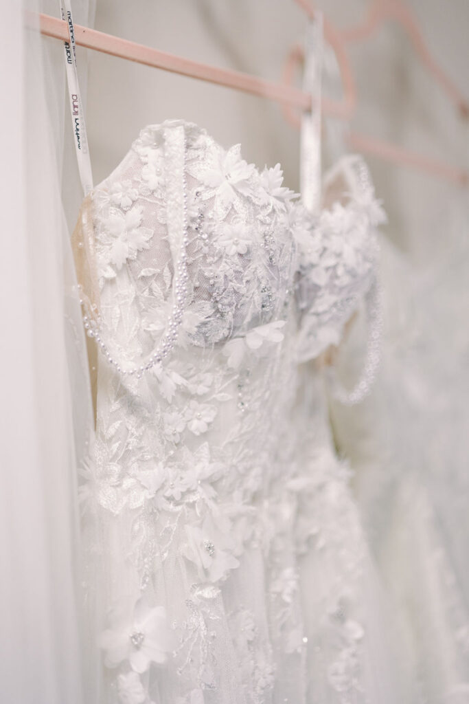 A white wedding dress with floral lace applique, bead details, and off-the-shoulder straps hangs on a pink hanger.