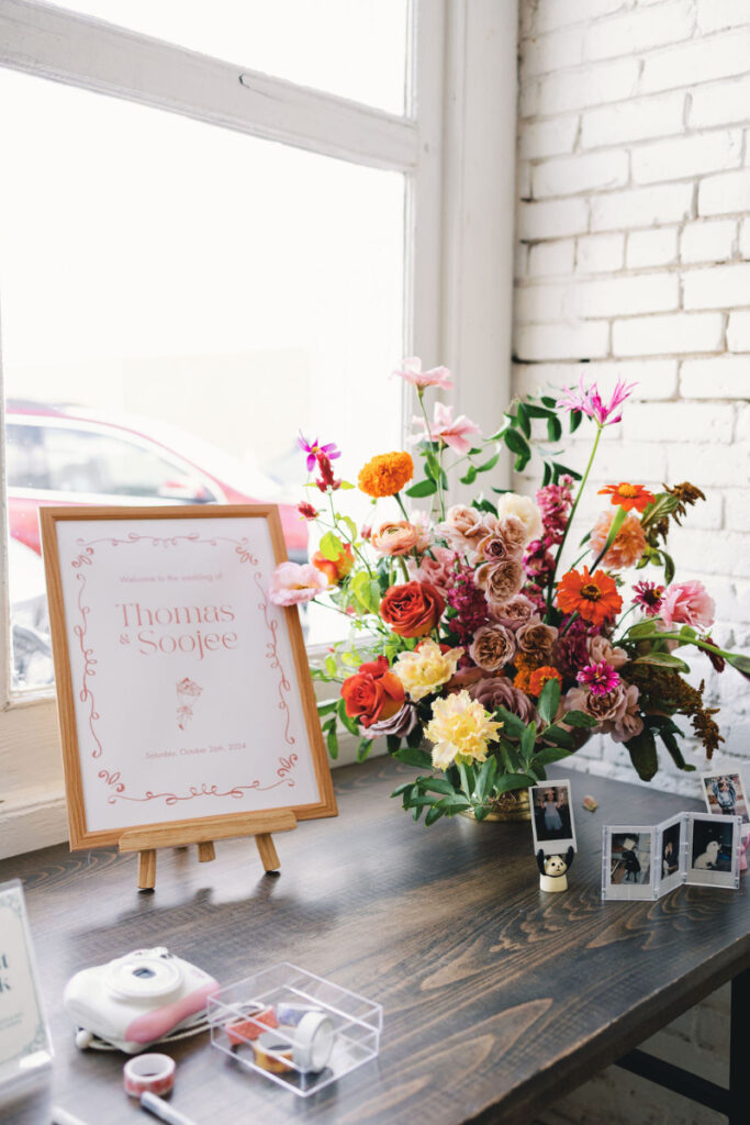 A framed sign with "Thomas & Soojee" and wedding date, a colorful floral arrangement, instant camera, film, and small photos displayed on a wooden table by a window.