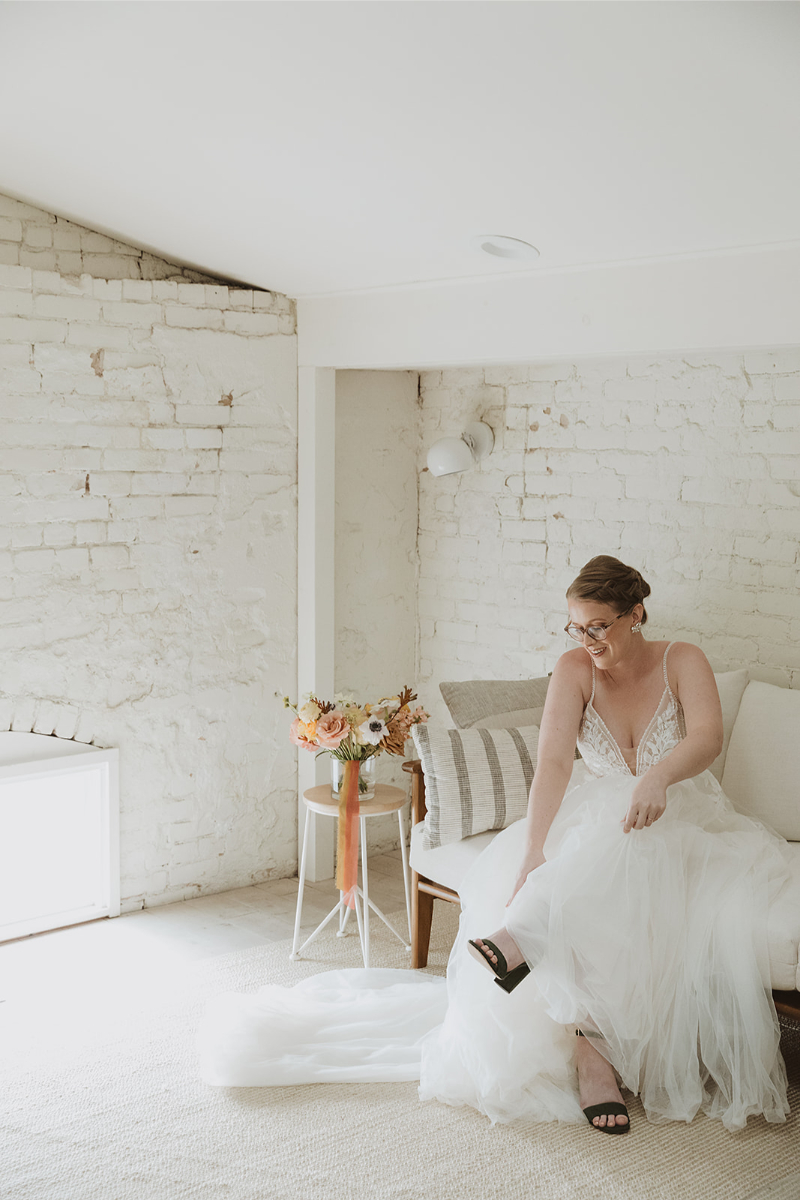 A bride in a white gown sits on a couch, adjusting her shoe. A bouquet of flowers is on a small table beside her in a bright room with white brick walls.