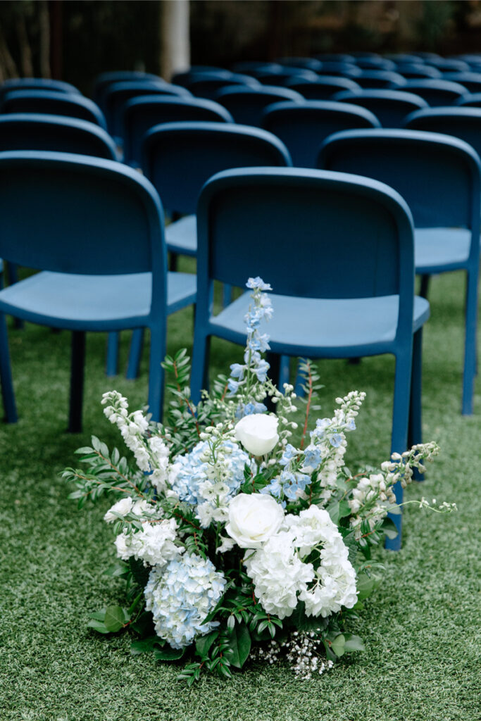 Rows of blue chairs on grass with a floral arrangement of white and light blue flowers in the foreground.