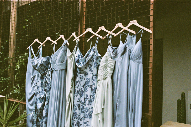 Six light blue and floral dresses on hangers are displayed outside on a wooden rack against a wire mesh background with some greenery visible.