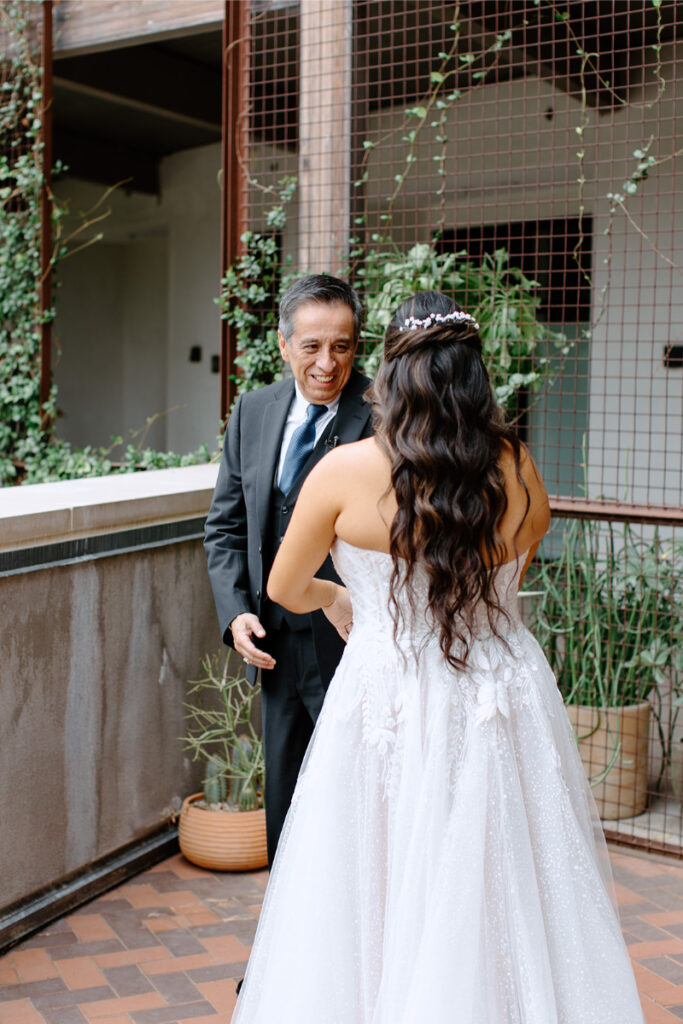 A man in a suit smiles at a woman in a white wedding dress with long wavy hair, standing on a tiled patio with potted plants and a metal grid in the background.