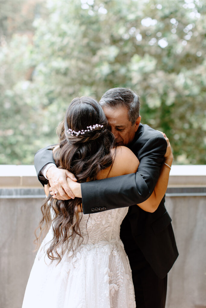 A bride in a white dress hugs an older man in a suit outdoors, both with eyes closed, surrounded by greenery.