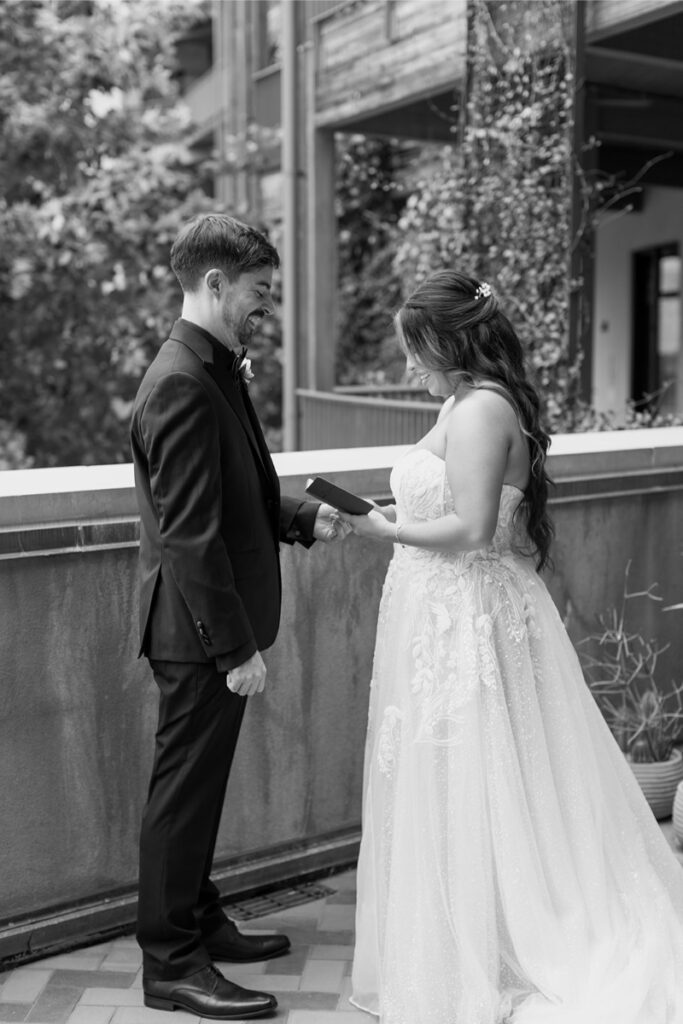 A bride and groom stand facing each other outdoors, smiling as the bride hands the groom a card. The scene is in black and white.