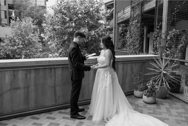 A bride and groom stand on an outdoor patio, holding hands and facing each other as the groom reads from a piece of paper.